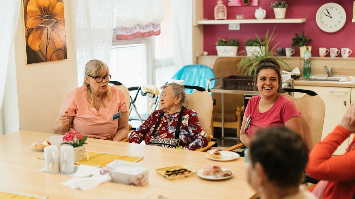 Elderly residents and caregivers enjoying a meal and conversation in a cheerful dining space.